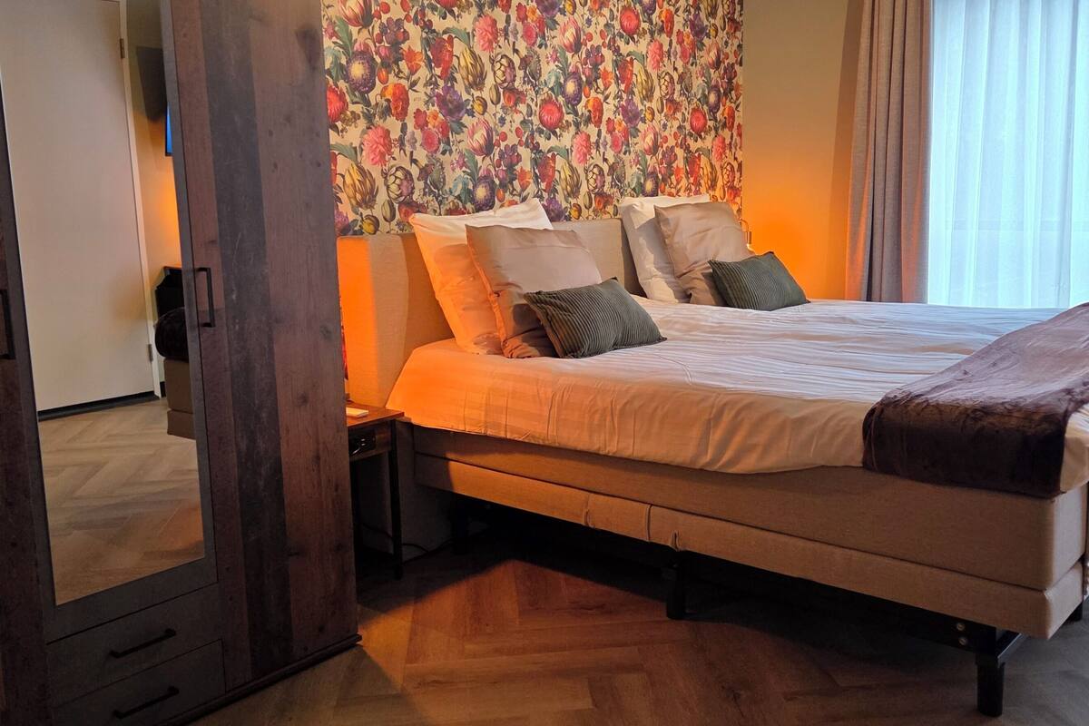 Un dormitorio con cama, piso de madera, papel tapiz floral y una ventana con cortinas.