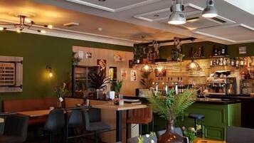A restaurant interior with wooden tables, chairs, hanging pendant lights, and a bar area with shelves of bottles.