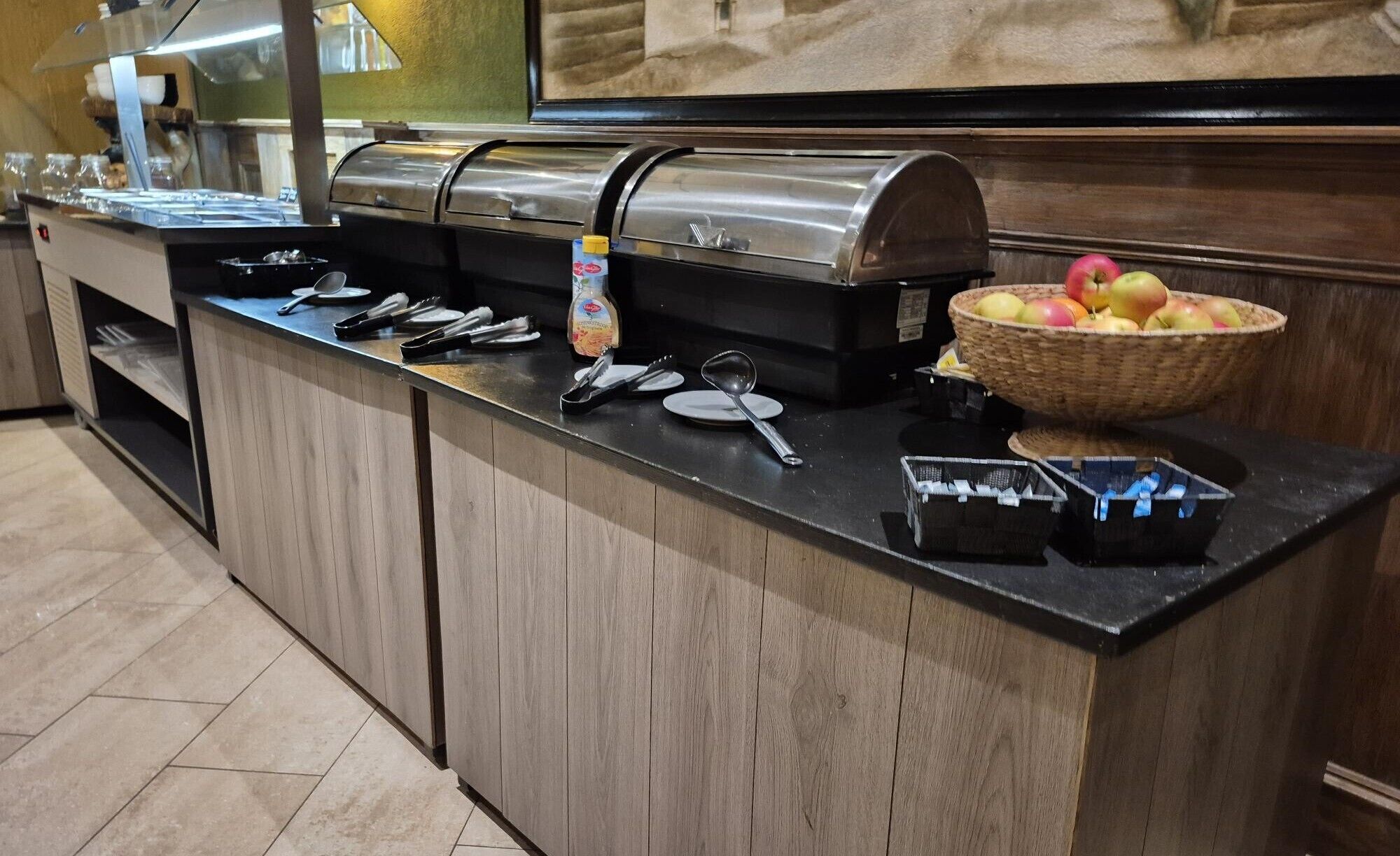 A buffet counter with a stainless steel food warmer, serving utensils, a bowl of fruit, and a bottle of milk.