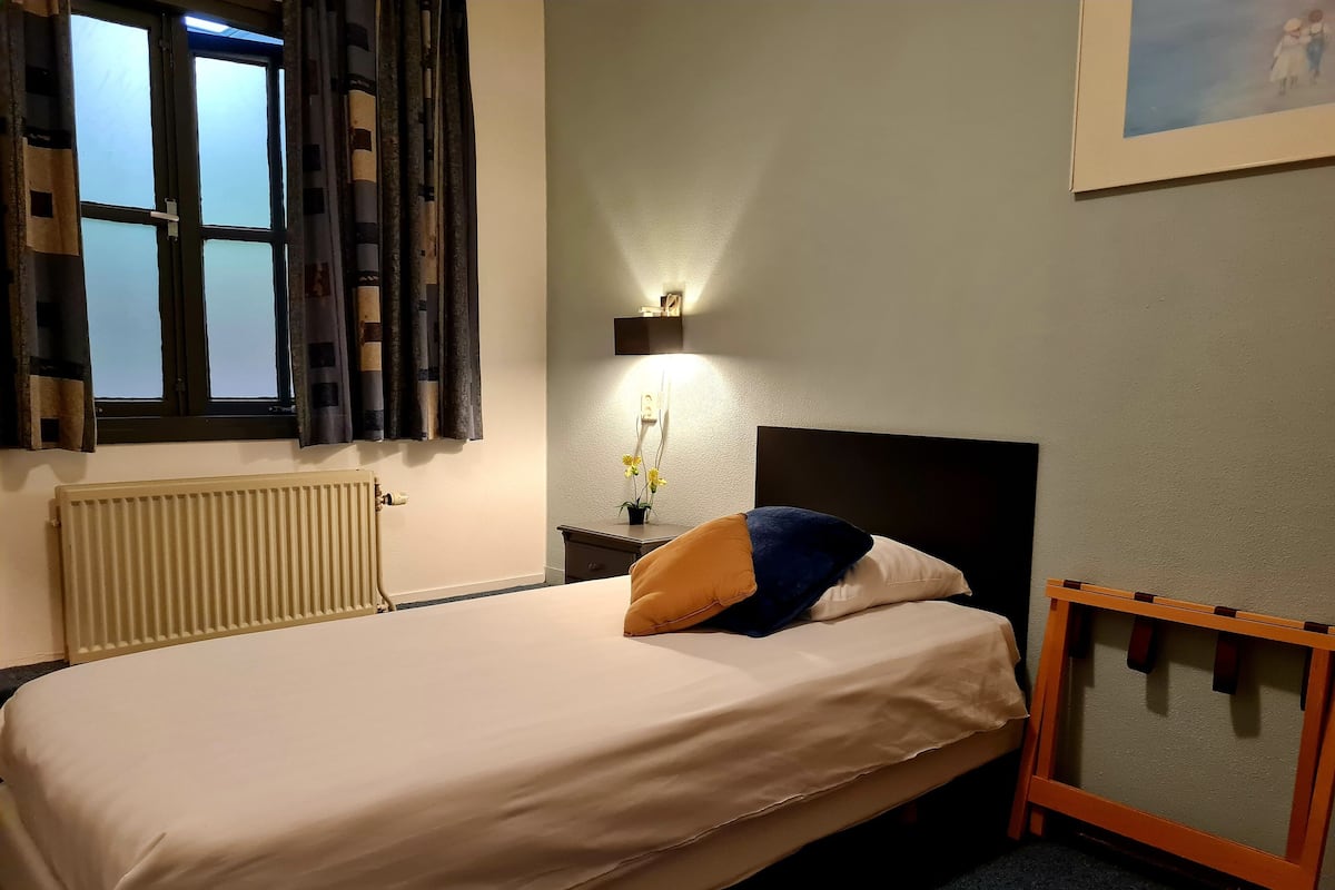 Una cama con cabecera, una mesita de noche con lámpara, un radiador y una ventana con cortinas.