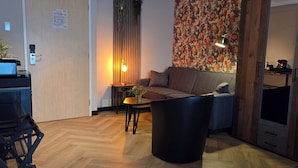 A modern living room with a sofa, a coffee table, a black trash bin, a wooden floor, a wall-mounted TV, and a door with a wooden frame. - Hotel 't Trefpunt (Made)