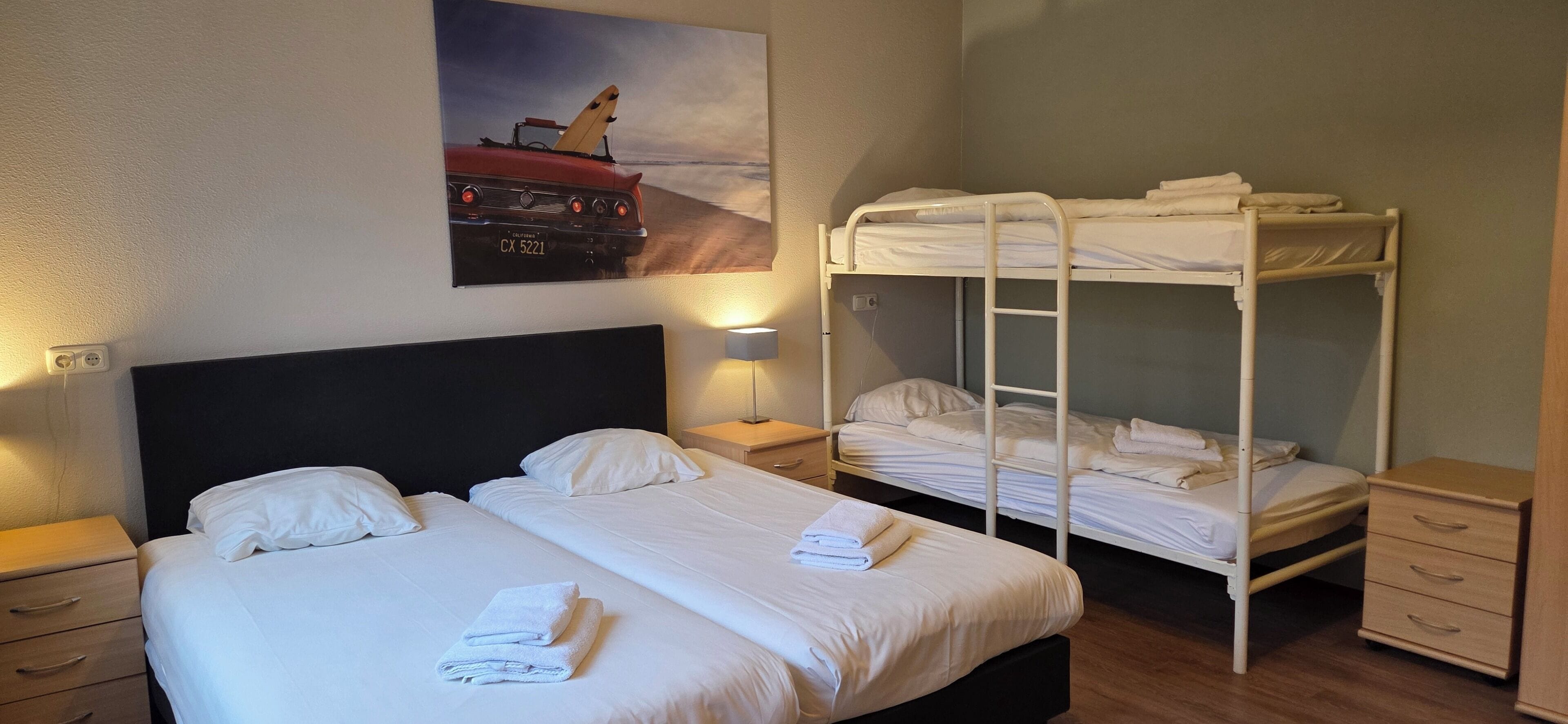 A hotel room with a bed, a bunk bed, a nightstand, a lamp, and a painting on the wall.