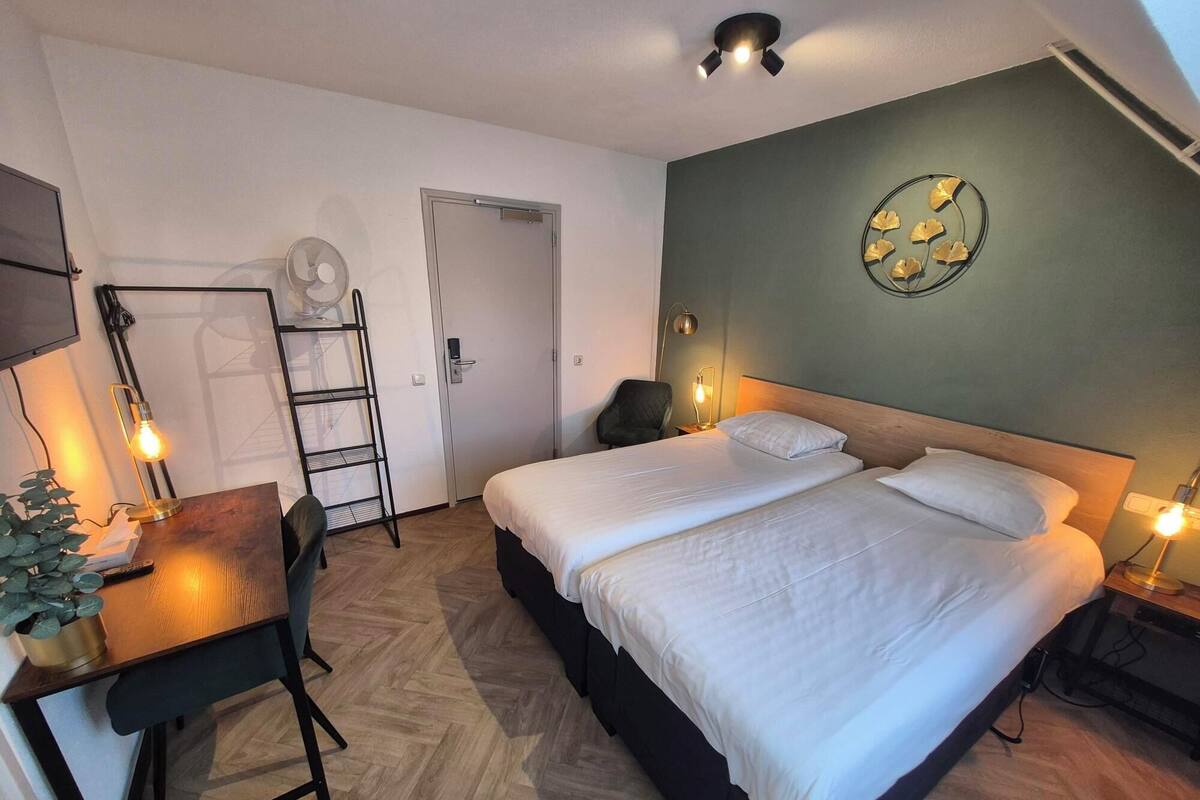 Habitación de hotel con cama, mesitas de noche, un escritorio con silla, armario, espejo y un reloj de pared.
