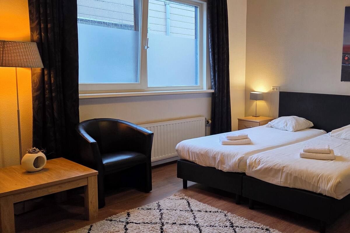 Una cama doble con sábanas blancas, una mesita de noche con lámpara, una silla, un radiador, una ventana con cortinas y un cuadro colgado en la pared.