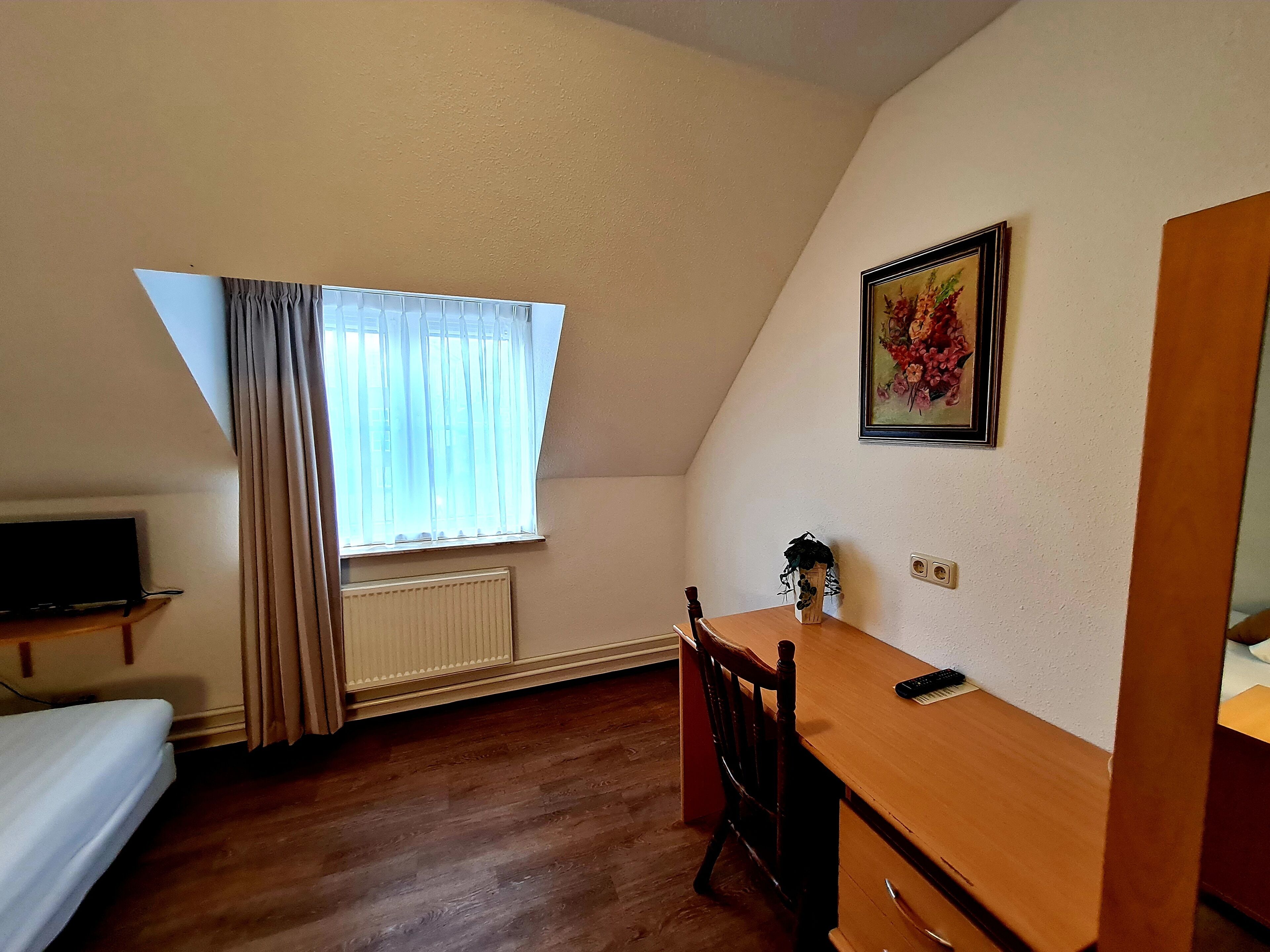Een kleine, driehoekige kamer met een houten bureau, een stoel, een bed en een raam met gordijnen.