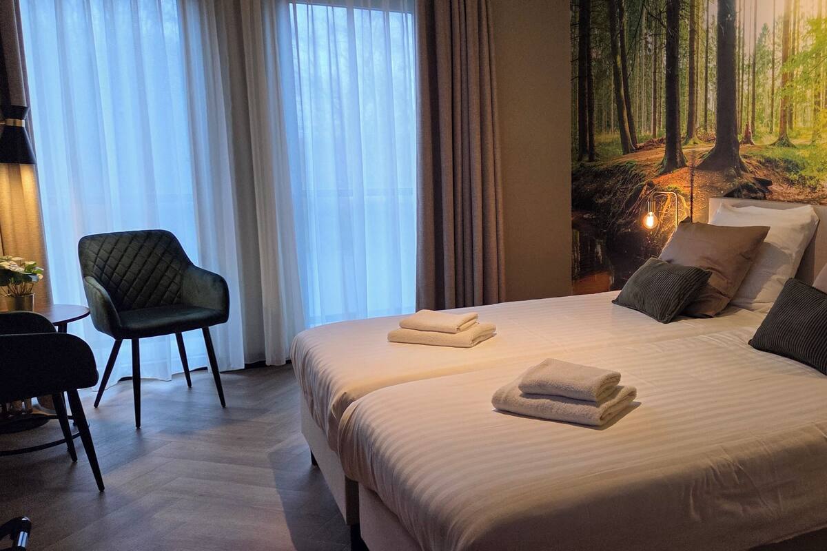 Una habitación de hotel con una cama grande, una silla, un cuadro de un bosque y una lámpara.