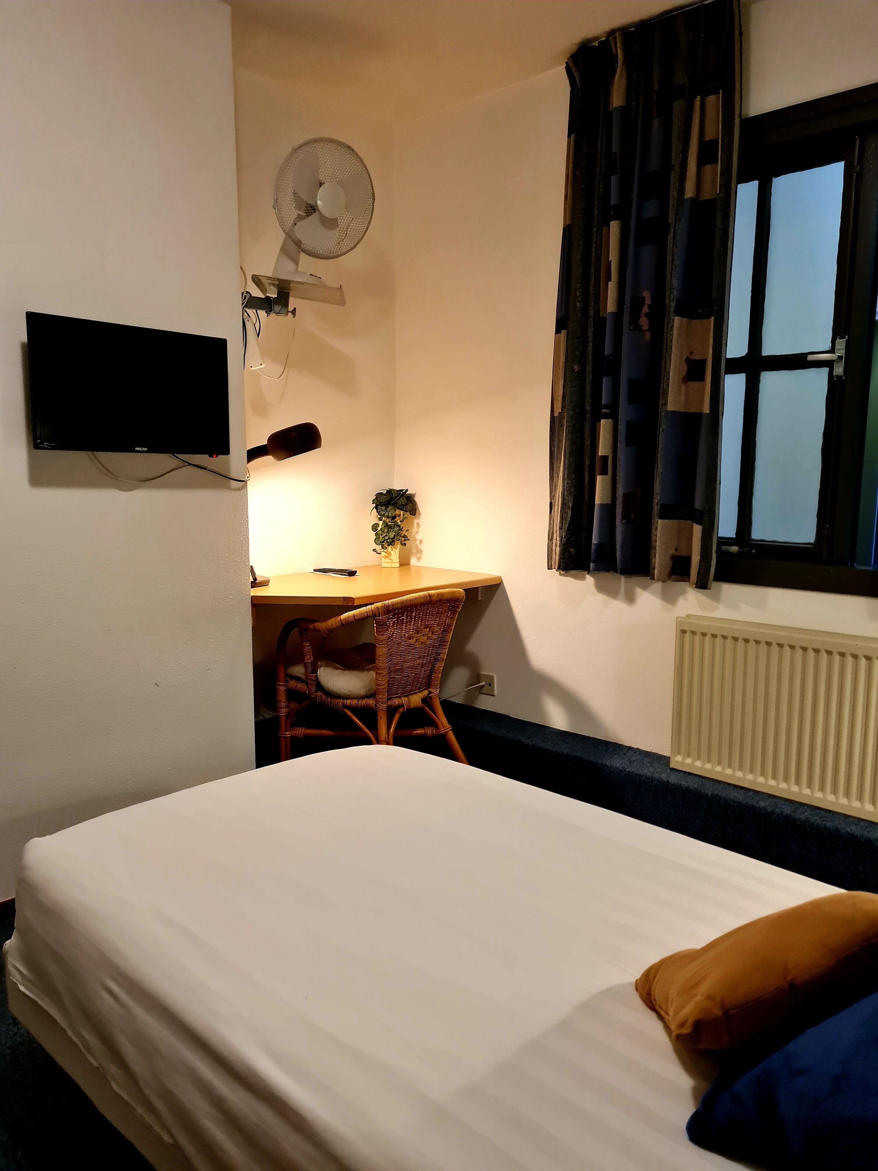 A bedroom with a bed, a desk with a chair, a wall-mounted TV, a fan, and a window with curtains.