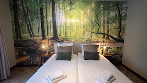 A room with two beds, a large forest mural, and a nightstand with a lamp. - Hotel 't Trefpunt (Made)
