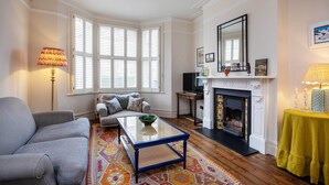 A living room with a sofa, a coffee table, a fireplace, and a large window with shutters. - Idyllic Elegance, by Veeve (London)