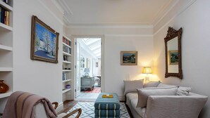 A well-lit living room with a sofa, ottoman, and armchair, a bookshelf, and a painting on the wall. - Idyllic Elegance, by Veeve (London)