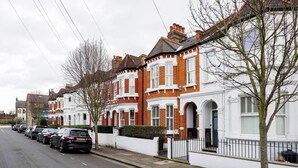 A row of red-brick houses with white trim and bay windows, parked cars along the street, and a tree in the foreground. - Idyllic Elegance, by Veeve (London)