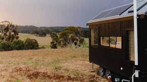 A small, dark-colored mobile home with a solar panel roof, situated on a grassy field with trees in the background.