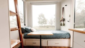 Habitación pequeña con cama, una mesita, una escalera y ventana con vista a árboles.
