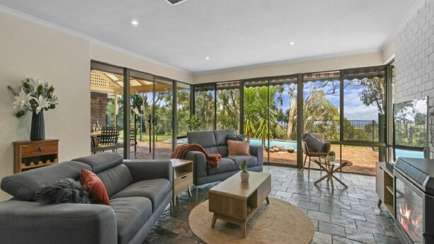 A spacious living room with a grey sofa, a coffee table, and a fireplace. Large windows offer a view of a pool and outdoor area. - Southern Vales by Wine Coast Holiday Rentals (WILLUNGA SOUTH)