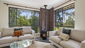 A cozy living room with two sofas, a glass coffee table, and a fireplace. Large windows offer a view of greenery outside. - Southern Vales by Wine Coast Holiday Rentals (WILLUNGA SOUTH)