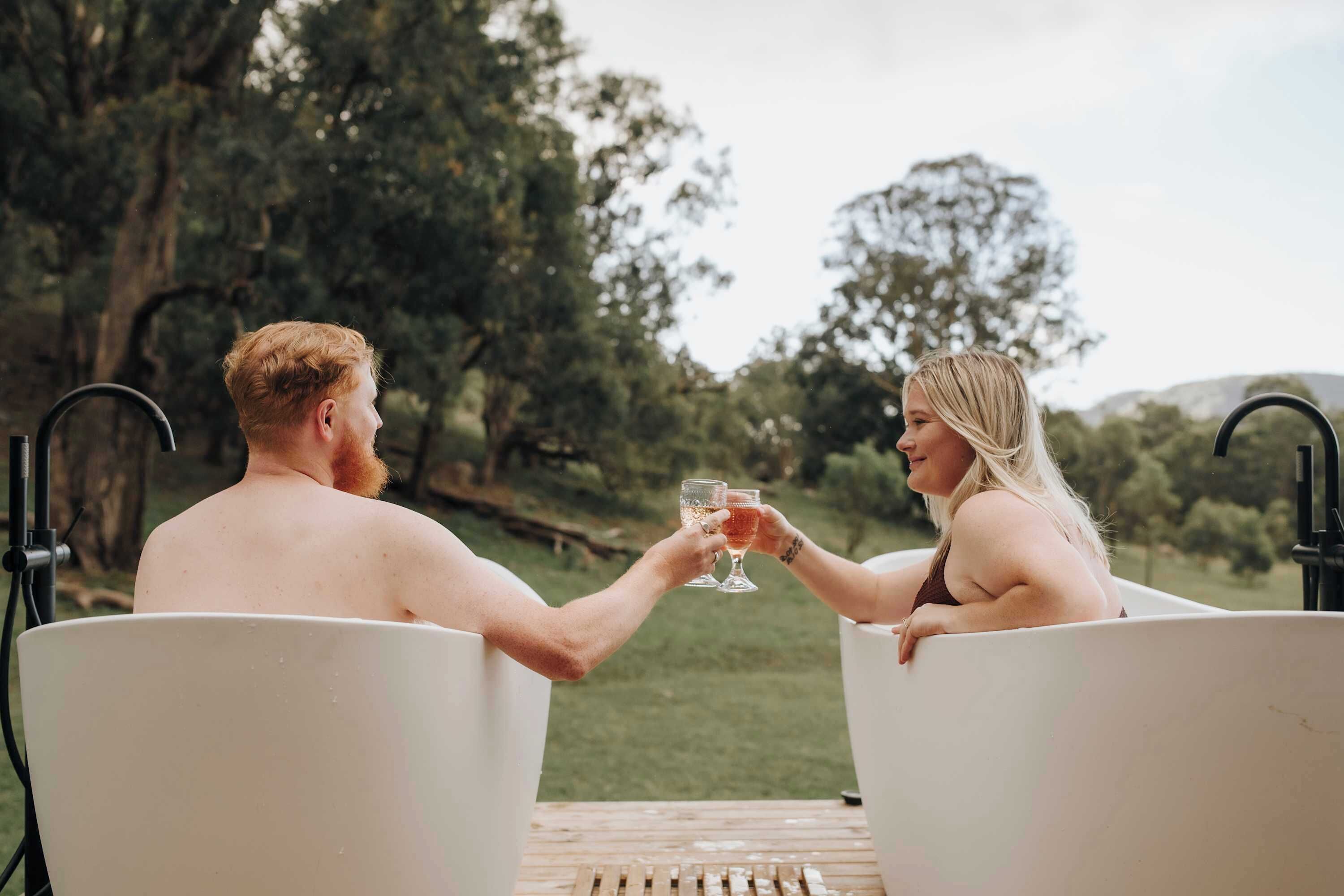 Two people sitting in white tubs outdoors, toasting with drinks.