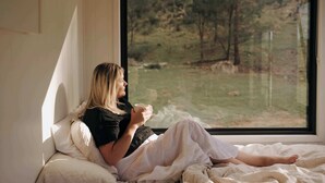 A person sitting on a bed with a window, holding a cup.