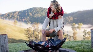A couple embracing by a fire pit in a grassy outdoor setting with hills in the background. - Tiny Mowgli Summary: (Bulliac)