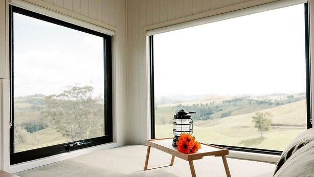 A room with large windows offering a view of rolling hills and trees. A small wooden table with a lantern and flowers is placed near the window. - Tiny Mowgli Summary: (Bulliac)