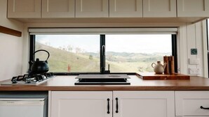 A kitchen with a view of rolling hills, a kettle, a stove, and a wooden cutting board. - Tiny Mowgli Summary: (Bulliac)