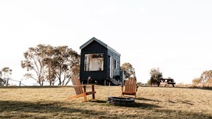 A small, dark-colored cabin with a pitched roof, two wooden chairs, and a picnic table in a grassy field with trees in the background. - Tiny Ruby - Central West (Duramana)
