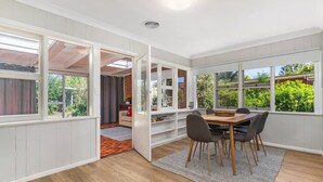 A room with a dining table, chairs, a bookshelf, and large windows. - Small Villages Retreat - Escape the City Life (Blayney)