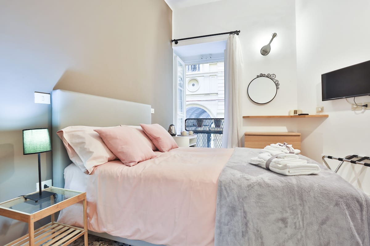 Un dormitorio moderno con cama, mesita de noche, lámpara y vistas de la ciudad a través de una ventana abierta.