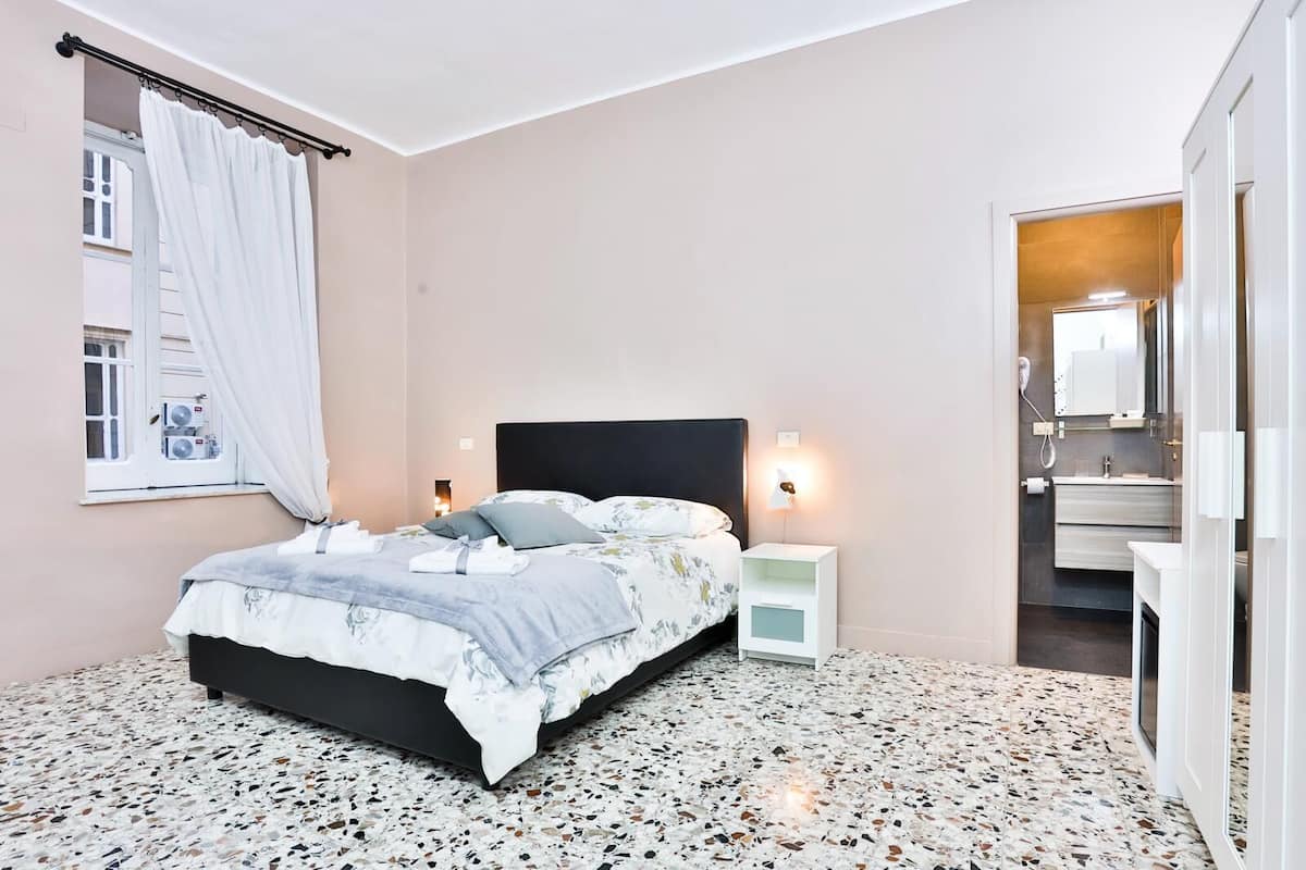 Un dormitorio con cama, mesitas de noche, una ventana con cortinas, un baño con lavamanos y espejo, y suelo de terrazo.