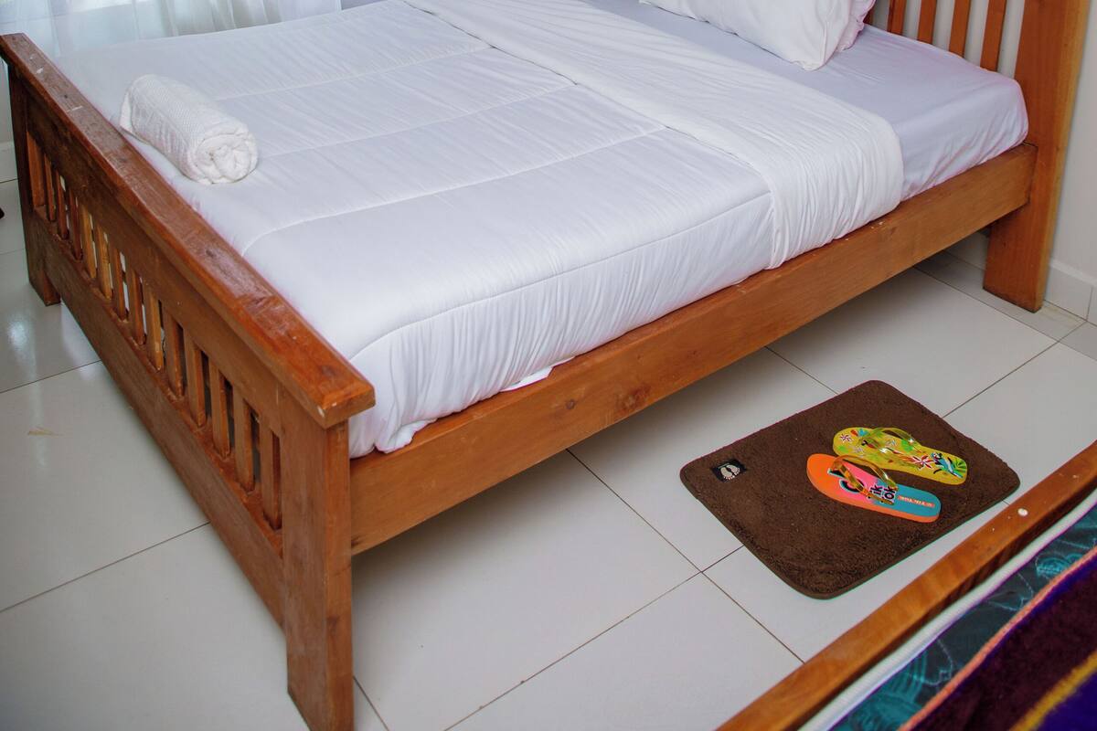 A neatly made bed with a wooden headboard, a white bedspread, and a pair of slippers on a brown mat.