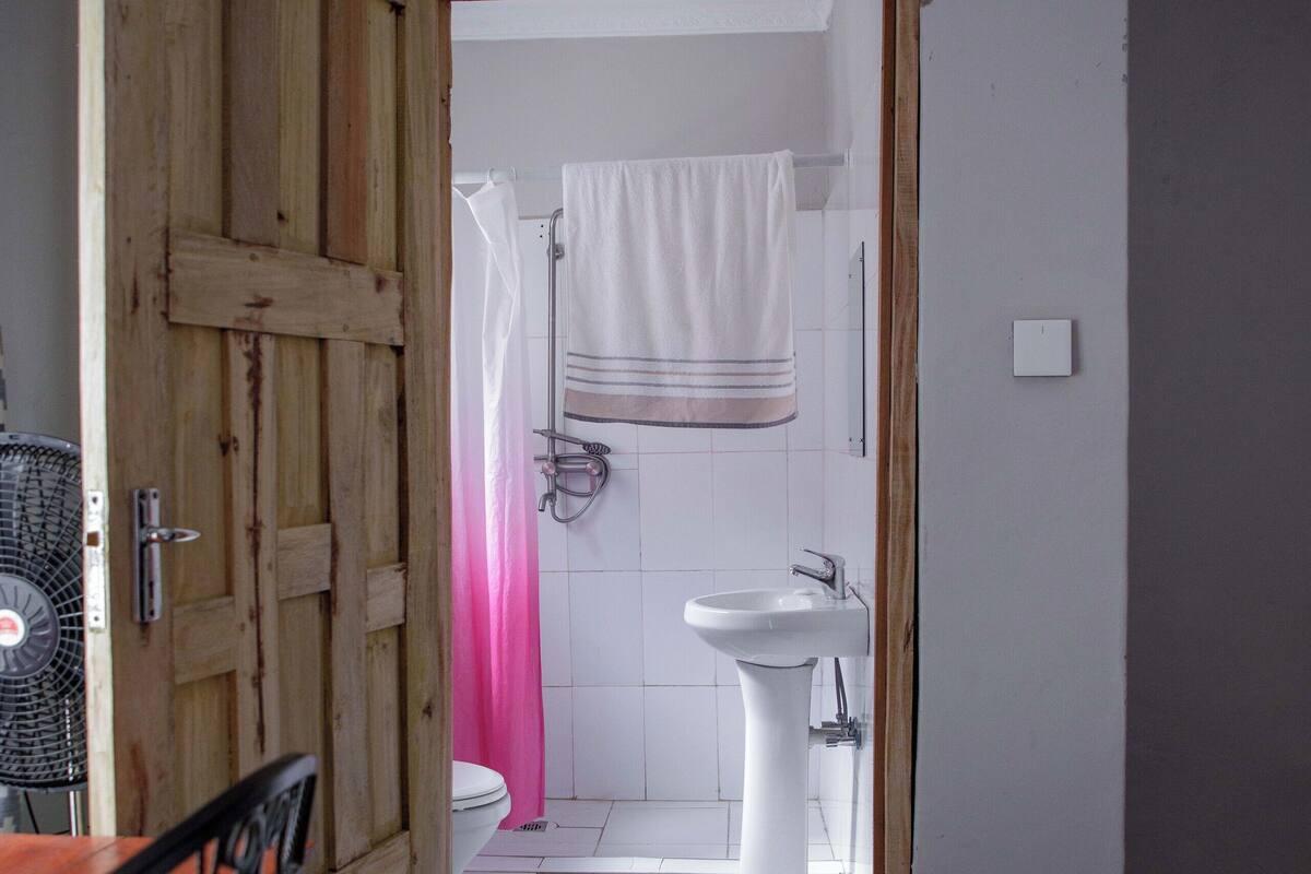 A room with a wooden door leading to a bathroom. The bathroom has a white sink, a toilet, and a shower with a pink curtain. There is a bed with white bedding in the room.