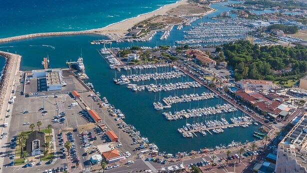 An aerial view of a marina with numerous boats docked, a beach in the background, and a mountainous landscape.