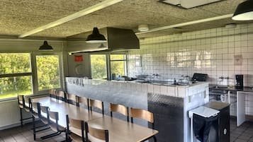 A restaurant with a long communal table, chairs, a kitchen area, and a view of greenery outside.
