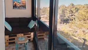 Un balcon avec un banc en bois et un canapé, une affiche accrochée au mur, et une vue dégagée sur les arbres et les bâtiments.