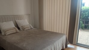 A bedroom with a bed, two pillows, a window with blinds, and a view of the outside. - Cozy Studio with Pool, Tennis, and Terrace - 500m from Beach (Argelès-sur-Mer)
