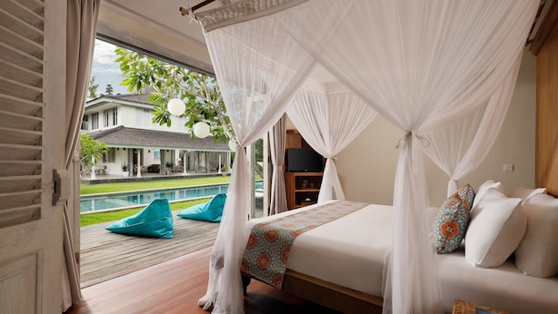 A bedroom with a bed, white canopy, teal bean bag chairs, and a view of a swimming pool and house. - Villa Hasian Jimbaran (Jimbaran)