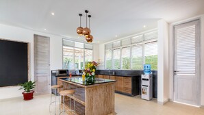 A modern kitchen with a central island, bar stools, a water dispenser, and a large window with blinds. - Villa Hasian Jimbaran (Jimbaran)