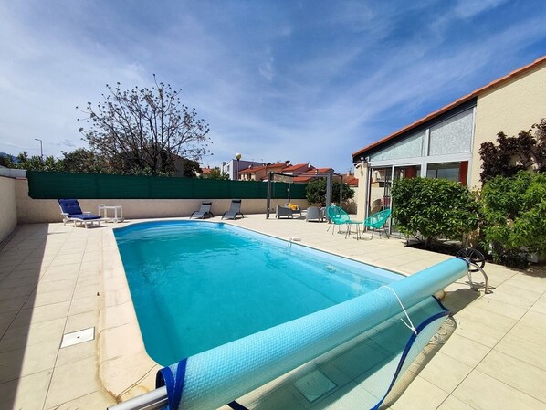 A rectangular swimming pool with a blue liner, surrounded by a tiled deck. There are several lounge chairs and a covered seating area with umbrellas.