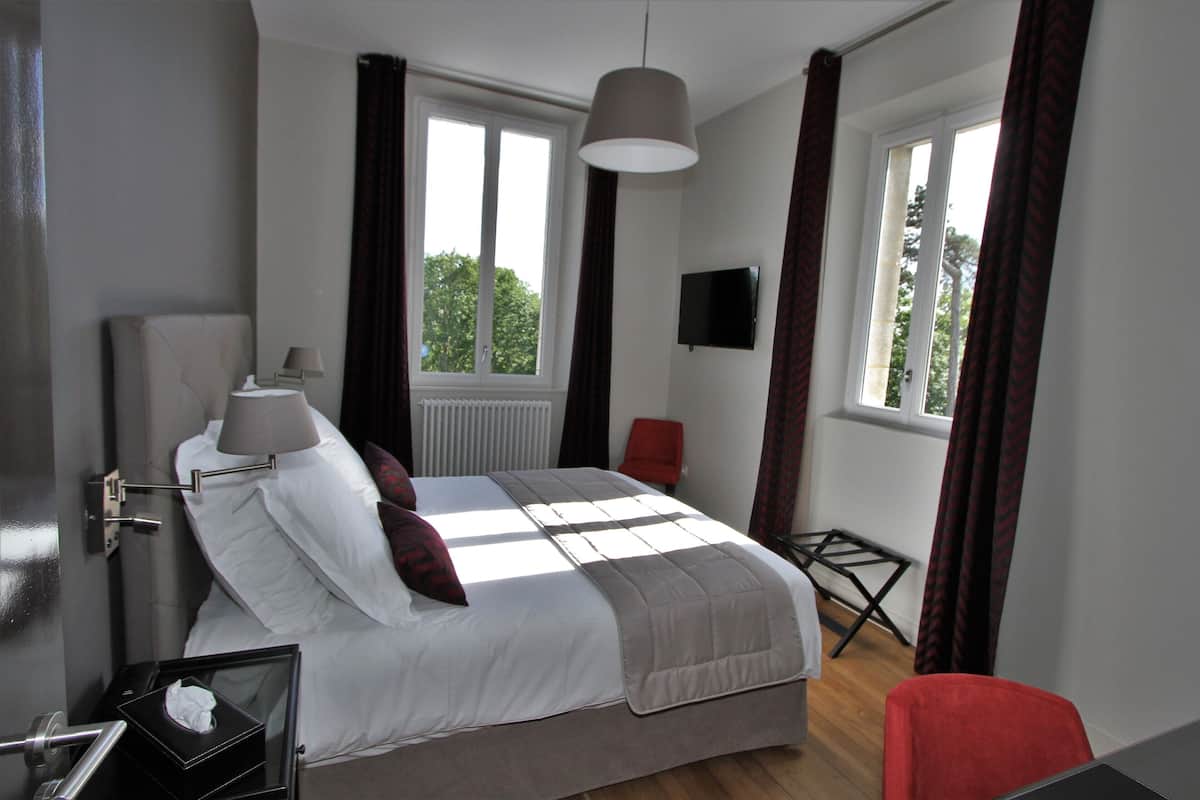 A bedroom with a bed, bedside table, lamp, TV, and windows.