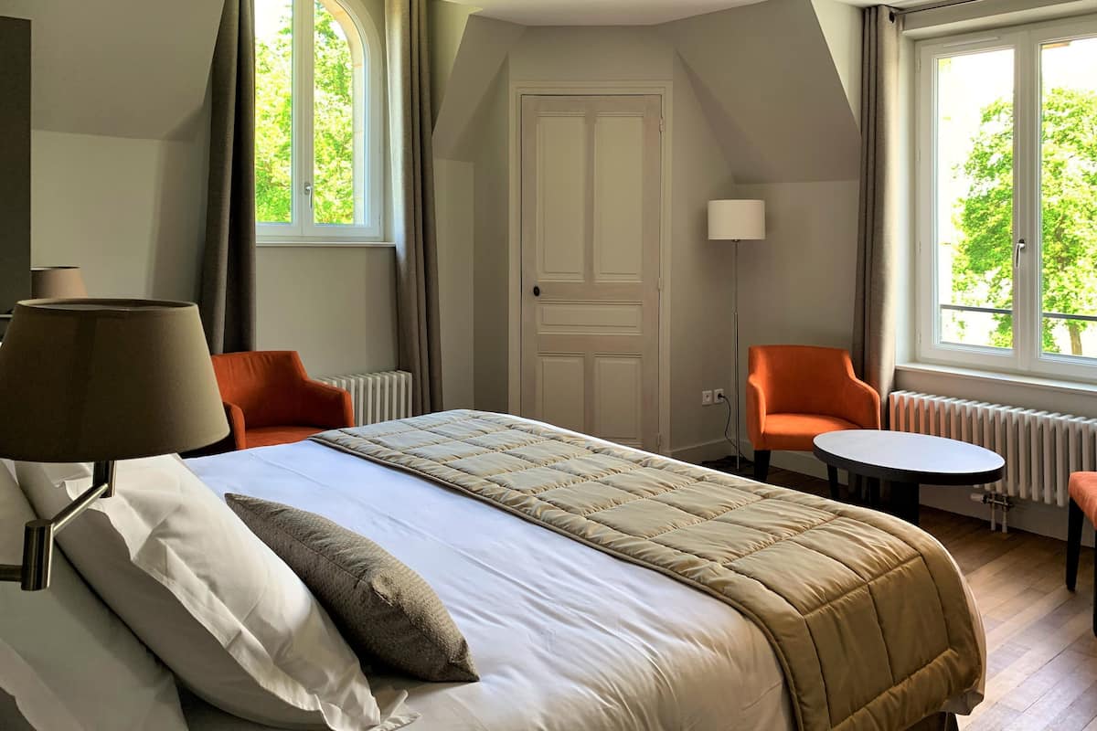 A hotel room with a bed, two chairs, a small table, a lamp, and a window with curtains.