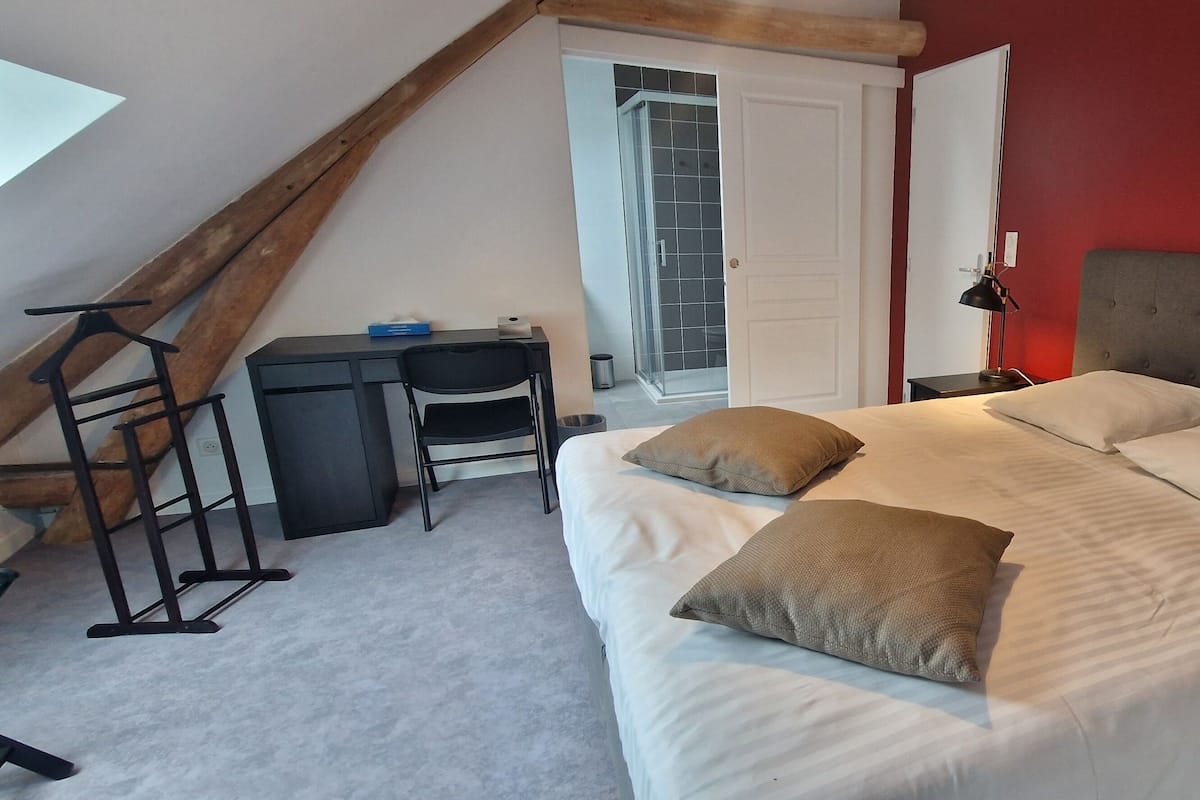 A bedroom with a bed, two pillows, a desk with a chair, and a window.