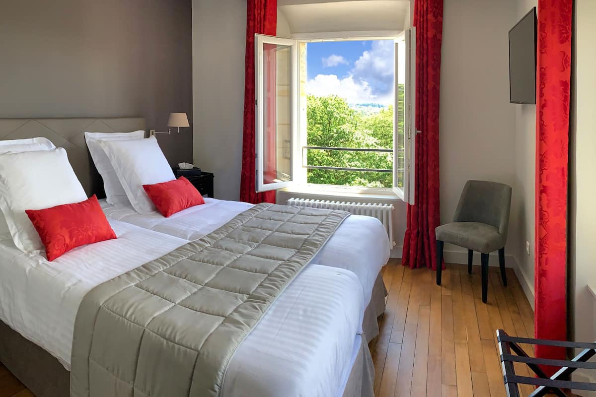 A hotel room with a large bed, red pillows, a chair, and a window with a view of greenery.