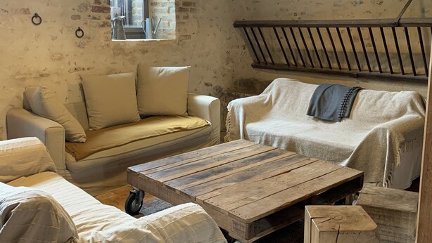 Una habitación rústica con paredes de ladrillo visto, techo de madera y un amplio ventanal. Hay dos sofás y una mesa de madera con ruedas.