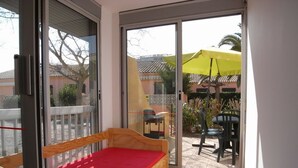 A small balcony with a wooden bench and a red cushion, a sliding glass door, and a view of a patio area with outdoor furniture and a yellow umbrella. - Ground Floor 2-Room Apartment Near Beach & Shops, Sleeps 4 (Argelès-sur-Mer)