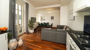 A modern living room with a grey sofa, a dining area with green chairs, a kitchen with a stove, and a large window with curtains.