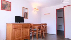 Une pièce comprenant un meuble en bois, un téléviseur, une table à manger avec des chaises, un miroir et une lampe fixée au mur.