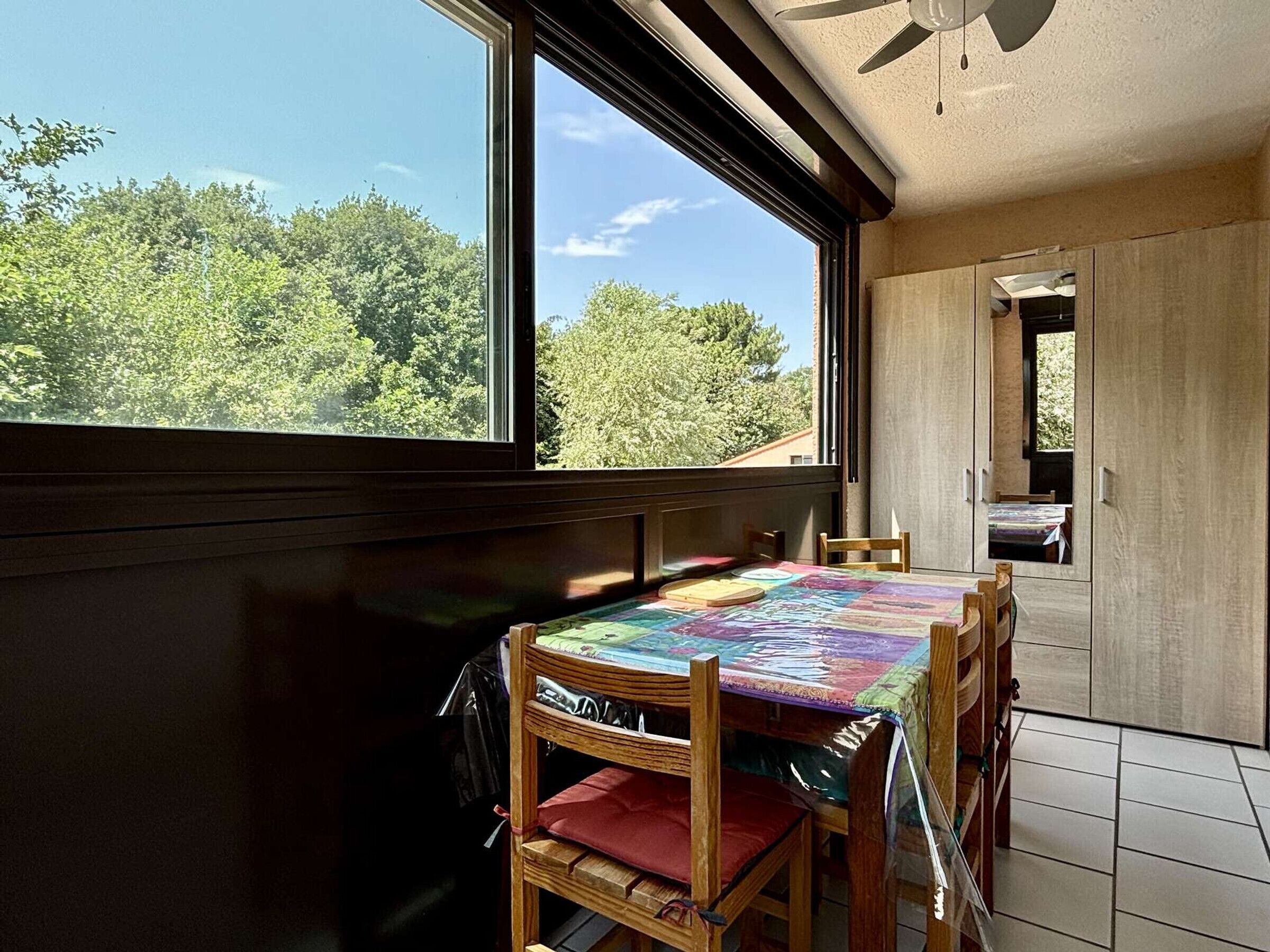 A dining area with a table covered by a colorful tablecloth, wooden chairs, and a view of trees through a large window.