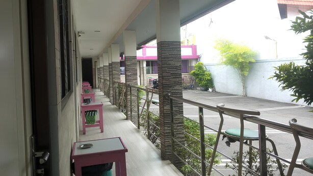 A covered outdoor area with a row of chairs and tables, a small trash bin, and a view of the street and buildings in the background. - Hotel Orchid Syariah (Tegal)