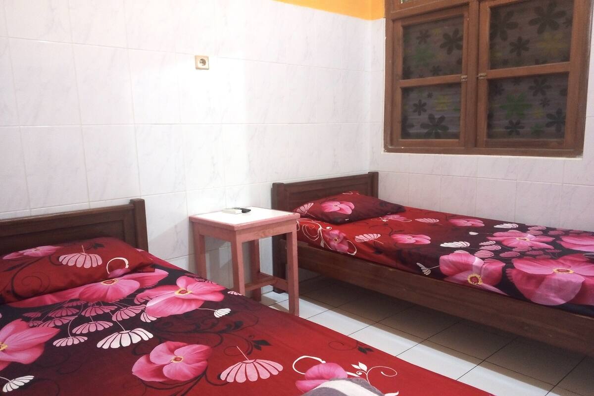 A room with two wooden beds with floral bedding, a small wooden table, and a window with floral curtains.