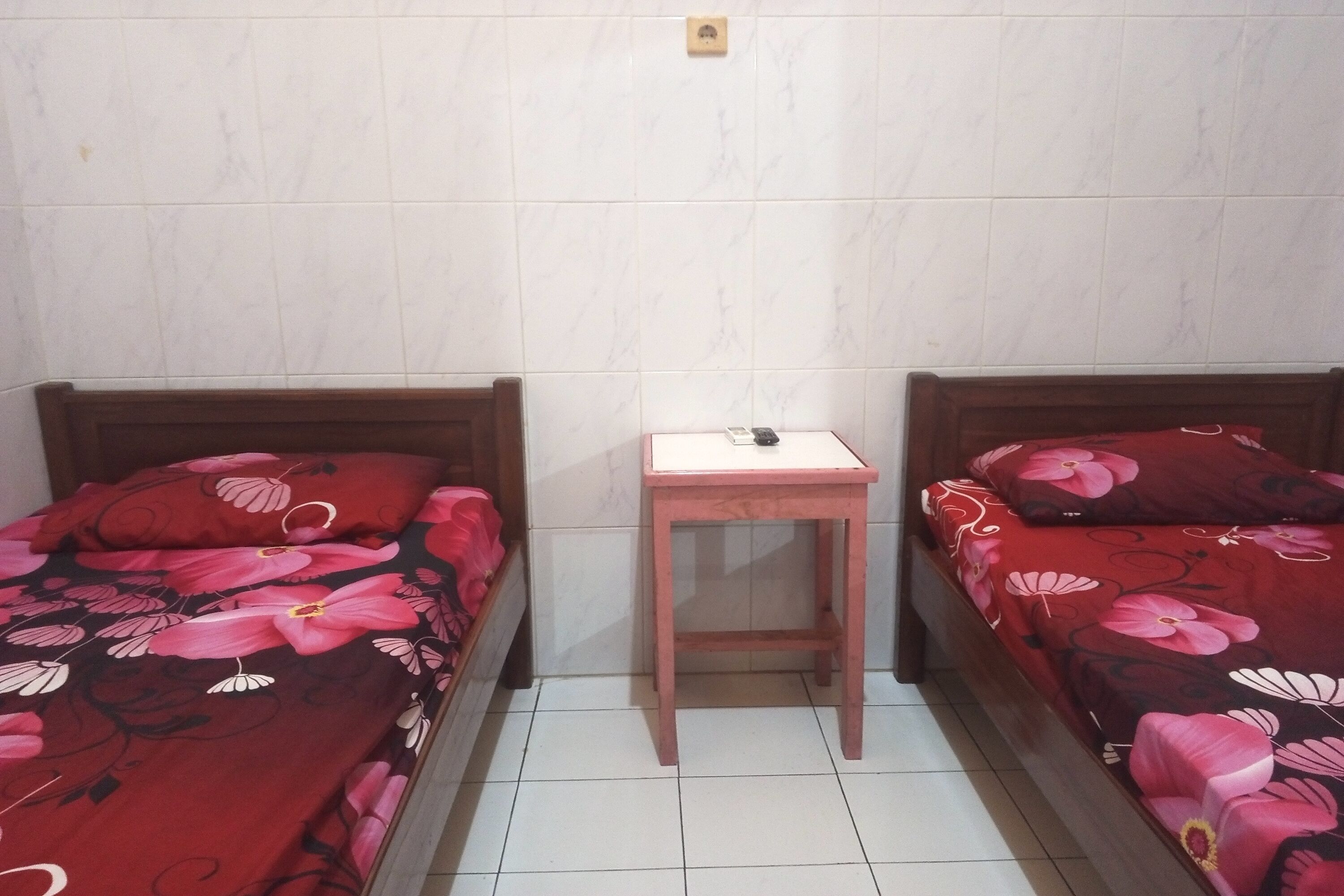 Een kleine kamer met twee eenpersoonsbedden, elk met een bloemenbedekking, een klein houten nachtkastje en een witte muur met een lichtschakelaar.