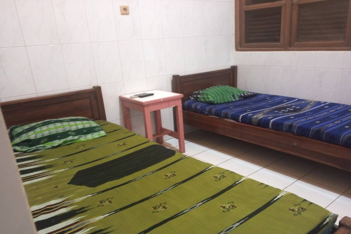 A small, single-room accommodation with two wooden beds, each with a distinct patterned bedspread. A small pink table is positioned between the beds.